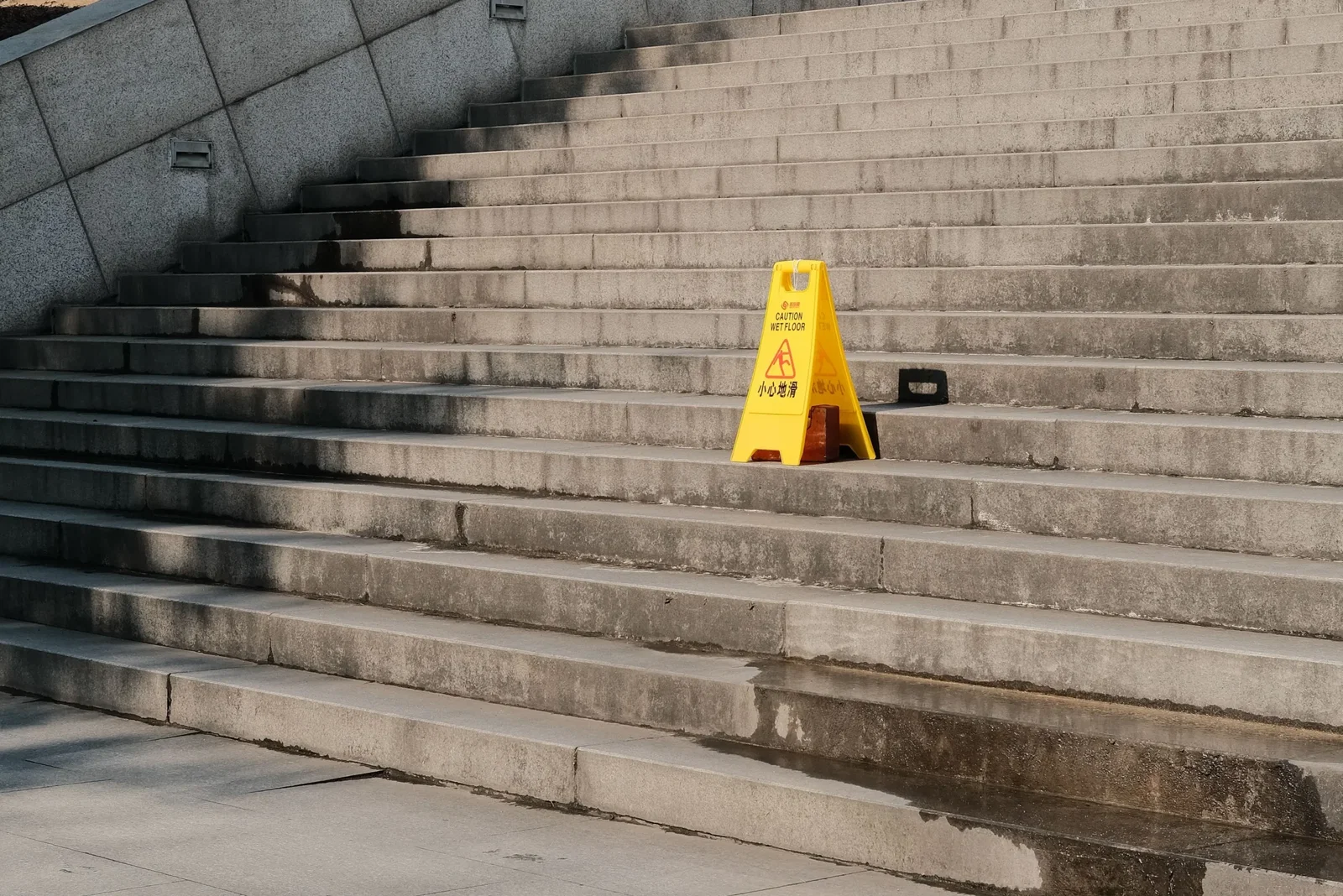 Concrete outdoor stairs with a bright yellow warning sign reading'Caution Wet Floor' on the lower steps; the ground is wet with reflections from puddles.